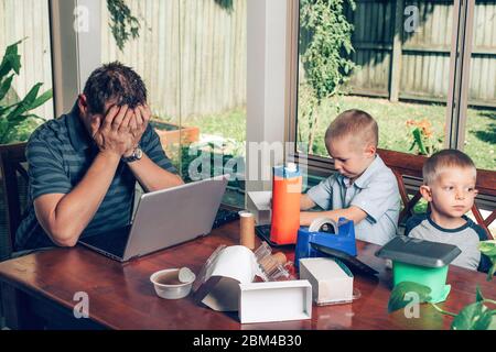 Concept of work from home and home schooling. Father trying to work on the Internet on a laptop while kids making crafts from recycled materials. Stock Photo