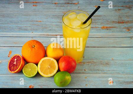 A tall glass cup of citrus juice with ice and a straw, stands on a table in a pile of whole and chopped oranges, lemons, lime and a small grapefruit. Stock Photo
