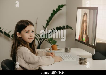 A young girl with long hair studying remotely from her female teacher online. A pretty child learns a lesson using a desktop computer at home. Home ed Stock Photo