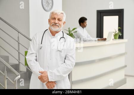 Portrait of eldery male doctor posing in white lab coat. Professional therapist with stethoscope on neck looking at camera, african man typing on lapt Stock Photo