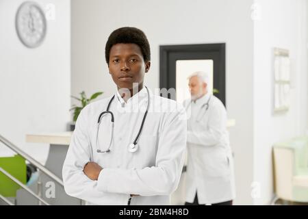 Portrait of african male doctor in white lab coat. Professional therapist with stethoscope on neck posing with arms crossed and looking at camera, eld Stock Photo
