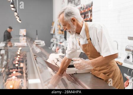 Side view of senior male butcher putting out of glass counter plate with sliced fresh steaks. Raw meat pieces in refrigerator with price tags ready for sale in meat department. Concept of food. Stock Photo