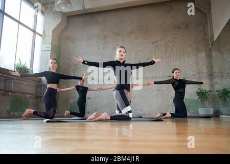 Side view of young fitnesswomen in sportswear practicing yoga pose, standing on knees with arms wide, practicing in studio, loft interior. Concept of healthy lifestyle, yoga. Stock Photo