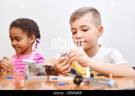 Front view of building kit for group of multiracial kids creating toys, having positive emotions and joy. Selective focus of lovely caucasian boy working on project, taking colorful parts. Stock Photo