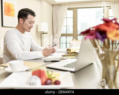 young asian man design professional working from home sitting at kitchen using cellphone (artwork in background digitally altered) Stock Photo