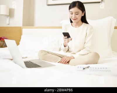 beautiful young asian woman sitting on bed working using mobile phone and laptop computer Stock Photo
