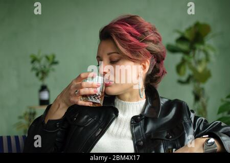 Young sad woman with dark pink hair with glam rock style look is drinking whisky from a glass on green background in studio, woman's alcoholism Stock Photo