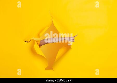 Female hand pointing, showing in torn yellow paper hole background. Breaking on, breakthrought with gesturing. Concept of celebration, shopping, proposal, sales, ad. Copyspace. Promotions, discounts. Stock Photo