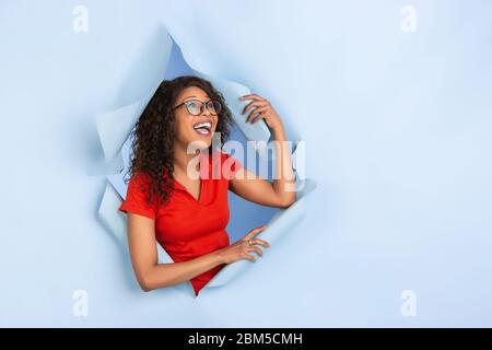 Wondered, shocked, greeting. Cheerful african-american woman in torn blue paper background, emotional, expressive. Breaking on, breakthrought. Concept of human emotions, facial expression, sales, ad. Stock Photo