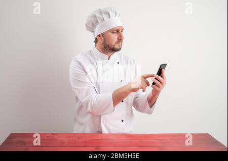 Portrait of young male chef in white uniform with blonde hair, holding smartphone, texting messages posing on a white isolated background Stock Photo