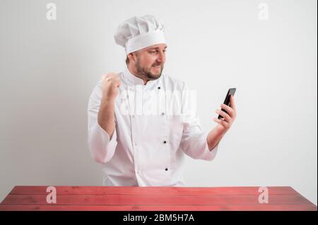 Portrait of young male chef in white uniform with blonde hair, holding smartphone, texting messages happy face posing on a white isolated background Stock Photo