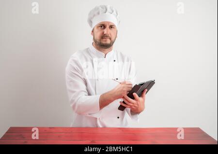 Portrait of young male chef in white uniform takes notes, looking at camera posing on a white isolated background