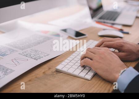 Cropped view of data analyst using computer keyboard near papers with graphs on table Stock Photo