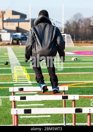 A high school boy is jumping over track hurdles with yellow mini hurdles and medicine balls in front of him for speed and agility practice. Stock Photo