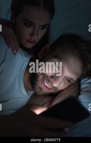 Angry woman embracing smiling boyfriend with smartphone on bed at night Stock Photo