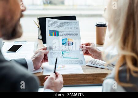 Selective focus of business people holding paper with charts near gadgets and paper cup on table Stock Photo