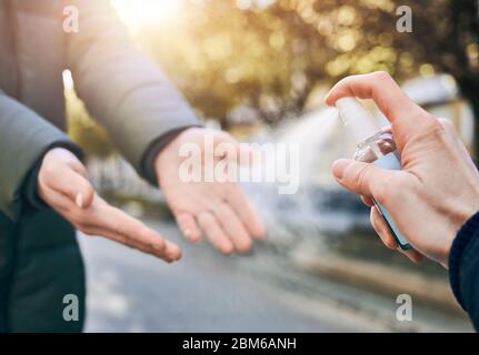 Selective focus on male's hand spraying sanitizer on woman's hands outdoors to disinfect hands, image full of sunlight. Stop coronavirus infection concept. Preventive measures during quarantine regime Stock Photo