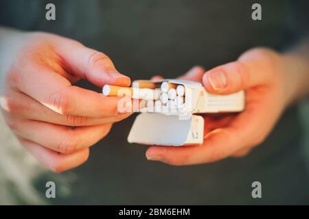 A young guy in a green t-shirt holds an open pack of cigarettes in his hands, and takes one out of it to light it. Stock Photo