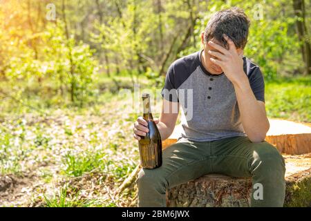 drunk sad man on a stump in the woods with a bottle of alcohol in his hand Stock Photo