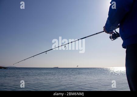 Unrecognizable male fisherman stands with a fishing spinning rod by the sea and waiting for fish on the right Stock Photo