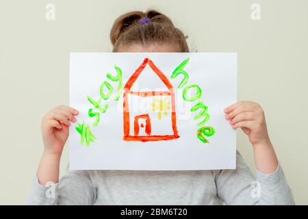 Child's hands holding a picture of red house and words Stay Home covering her face. Children in quarantine concept. Stock Photo