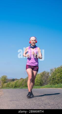 A young woman running on a country road on a sunny day dressed in black jogging shoes, dark pink shorts and purple tshirt. Stock Photo