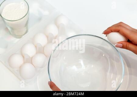 Close-up on the hands of a woman who cracks an egg with her fingers and pours the yolk into a plate against the background of a package with a dozen e Stock Photo