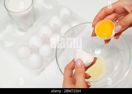 Close-up on the hands of a woman who cracks an egg with her fingers and pours the yolk into a plate against the background of a package with a dozen e Stock Photo