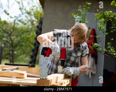 Senior Caucasian woman assembles a wooden frame of a building using metal fasteners using a screwdriver Stock Photo