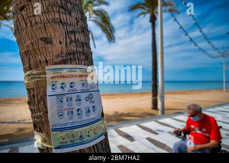 Benidorm, Alicante Spain, 4.5.2020, Corona crisis: man wearing face mask in wheelchair and Information on hygiene and safety on a poster on a tree on the promenade at Playa Levante Stock Photo