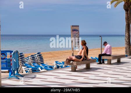 Benidorm, Alicante Spain, 4.5.2020, corona crisis: three people sit and lie on stone benches on the promenade on the deserted Playa Levante beach Stock Photo