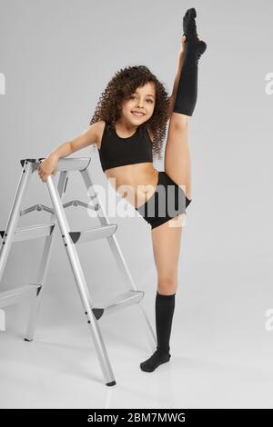 Portrait of adorable smiling girl in black sportswear and knee socks demonstraiting split while standing, isolated on gray background. Little female gymnast showing flexibility, leaning on staircase. Stock Photo