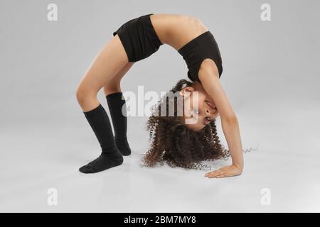 Cute smiling girl in sportswear and knee socks demonstraiting bridge exercise, isolated on gray studio background. Little female professional gymnast with curly hair showing flexibility, training. Stock Photo