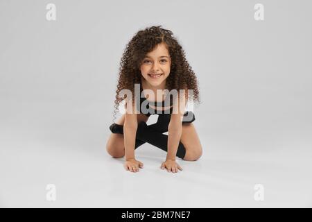 Close up of attractive smiling girl in black sportswear and long socks standing on knees in lotus pose of floor, isolated on gray studio background. Little professional female gymnast with curly hair. Stock Photo