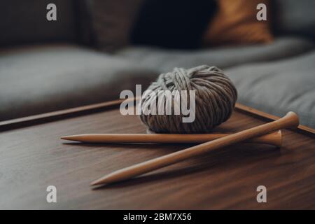Close up of a ball of grey chunky wool yarn and a pair of crossed large wooden knitting needles on a wooden table, shallow focus, in modern Scandi int Stock Photo
