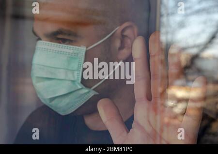 Infected caucasian man looking through the window in protective mask. Self isolation concept. Stock Photo