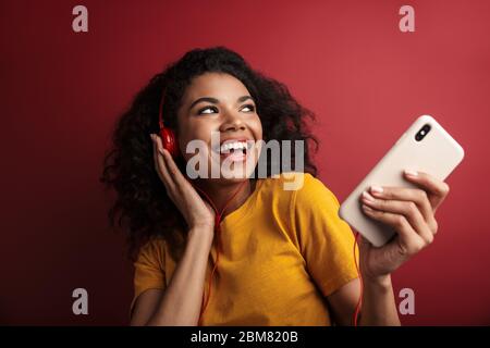 Image of beautiful brunette african american woman in headphones singing and holding cellphone isolated over red background Stock Photo