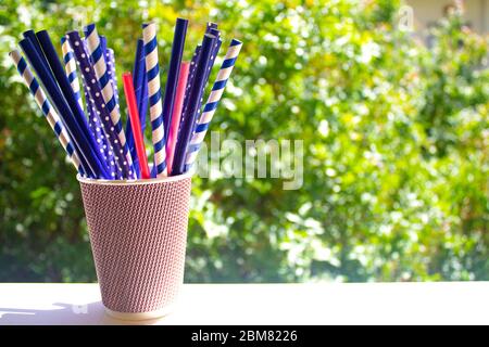 Several multicolored paper cocktail tubes in paper cup with green trees background. Ecologic saving. Stock Photo