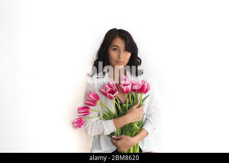 A portrait of charming dark haired young woman with many beautiful pink tulips on the white background. Stock Photo