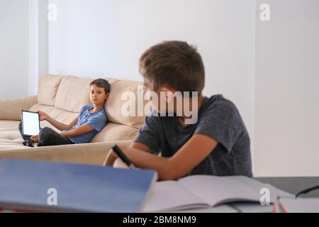 Brothers at home talking about homework tasks. Boy doing homework with books notebooks and other boy using digital tablet for online lessons Stock Photo