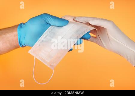 Man and woman hands in protective gloves holding medical face mask. On orange background Stock Photo