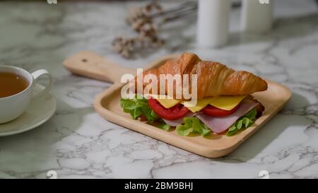 Close up view of breakfast meal with croissant sandwich ham, cheese on wooden plate and tea cup on marble desk Stock Photo
