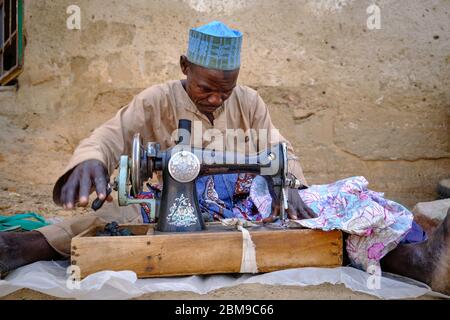 Tailor sitting on the floor sewing with an old machine at a market in the streets of Kiyawa. Stock Photo
