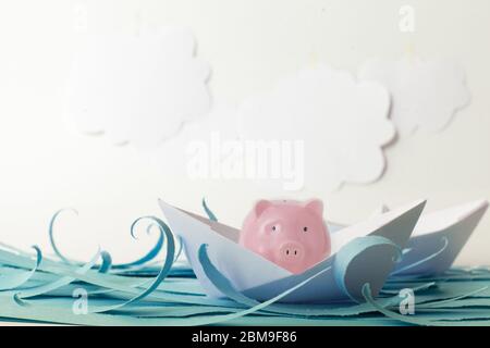 Pink piggy bank sits in a paper ship and floats on blue paper waves on background of white paper clouds. Concept of saving finance during a crisis. Ad Stock Photo