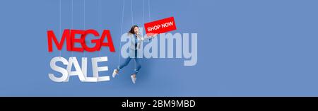 Excited beautiful Asian woman puppet on strings holding BUY NOW sign with MEGA SALE texts hanging on light blue banner background with copy space Stock Photo