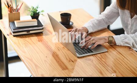 Cropped image of creative woman's hands typing on computer laptop that putting on wooden working desk and surrounded by stack of books, pencil holder, Stock Photo