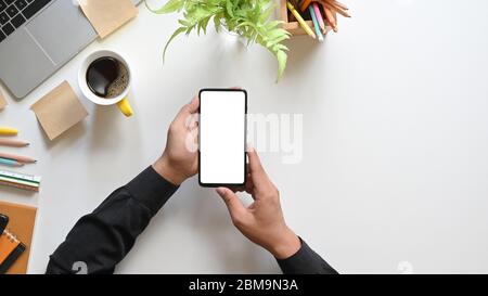 Top view hands holding a white blank screen smartphone over coffee cup, potted plant, pencils, computer laptop, notebook and ruler that putting togeth Stock Photo