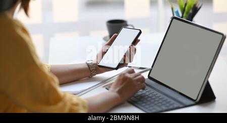 Cropped image of creative woman holding a smartphone and stylus pen while using a computer tablet with keyboard case while sitting at the white workin Stock Photo