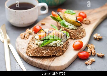 Bruschetta with grilled eggplant, cherry tomatoes, cheese dip, fresh basil and aromatic herbs on a wooden board. Healthy mediterranean cuisine. Stock Photo
