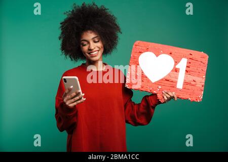 Image of african positive curly woman posing isolated over dark blue wall background holding blank with like symbol using mobile phone. Stock Photo